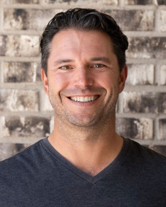 Dan Fernandez, smiling, in a charcoal v-neck in front of a brick wall.